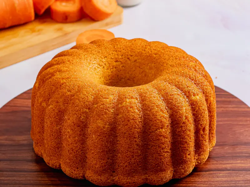 Bolo de cenoura caseiro em forma de anel sobre tábua de madeira, com cenouras ao fundo