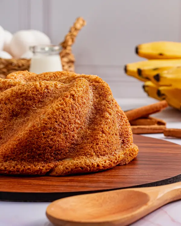 Bolo de banana em forma torcida, com bananas e canela ao fundo
