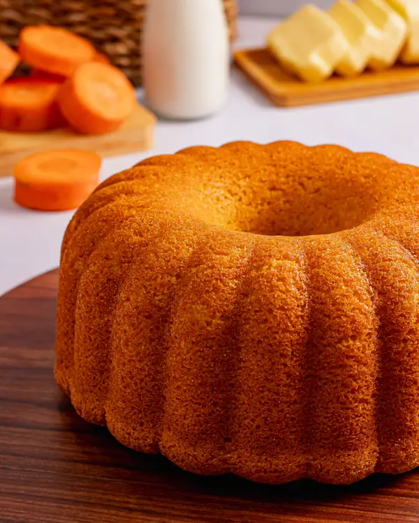 Bolo de cenoura em forma de anel sobre tábua de madeira, com cenouras fatiadas ao fundo